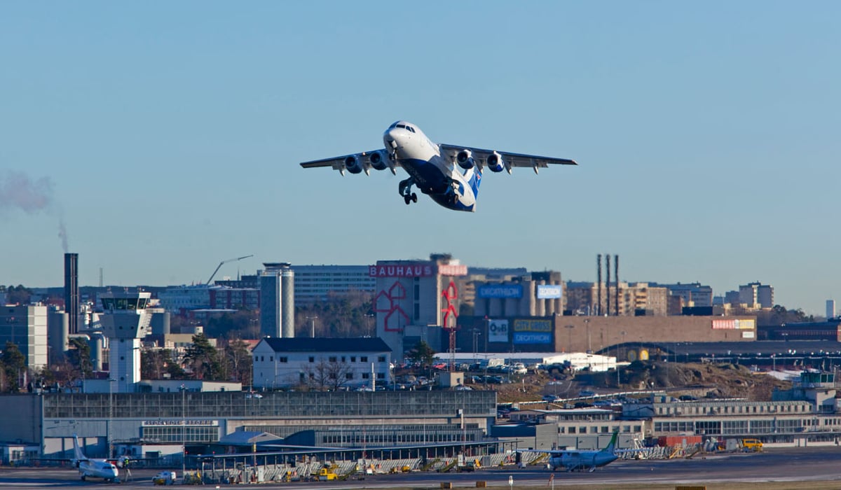 Flygplan på blå himmel ovanför flygplats, i bakgrunden visas olika varuhus