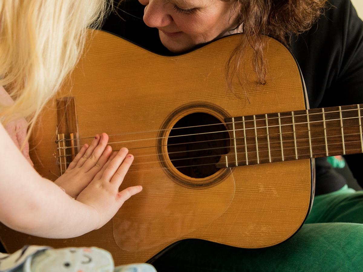 Foto på en vuxen kvinna som håller i en gitarr medan ett barn håller fram sina händer på gitarrens strängar och spelar.