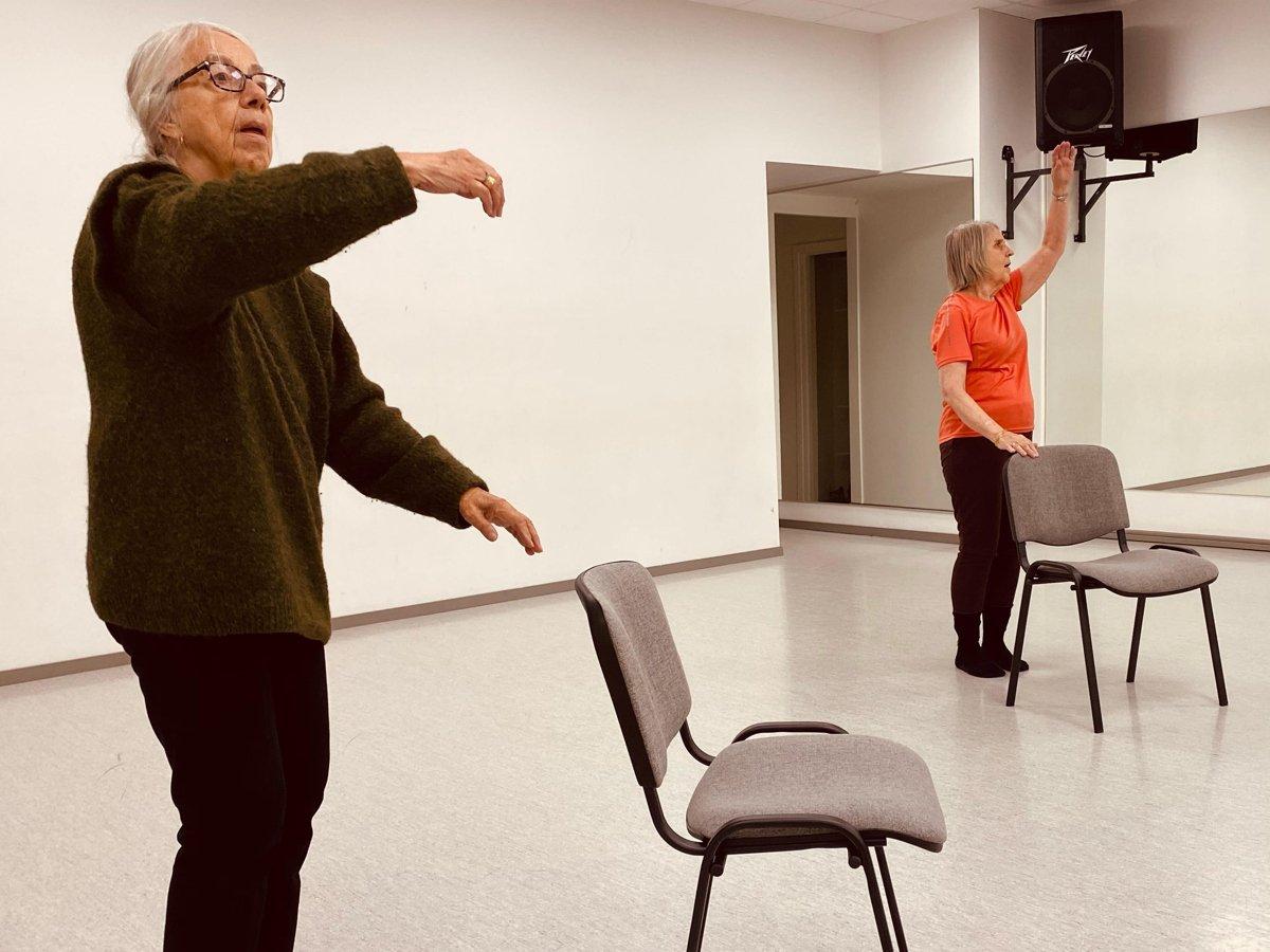 Två äldre kvinnor dansar bakom var sin stol.