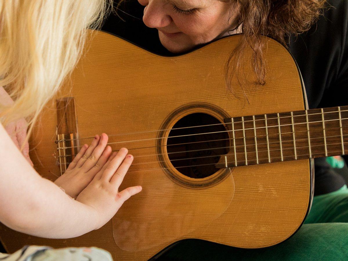 Foto på en vuxen kvinna som håller i en gitarr medan ett barn håller fram sina händer på gitarrens strängar och spelar.