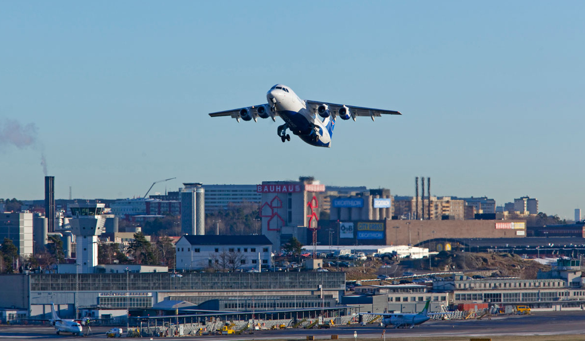 Flygplan på blå himmel ovanför flygplats, i bakgrunden visas olika varuhus