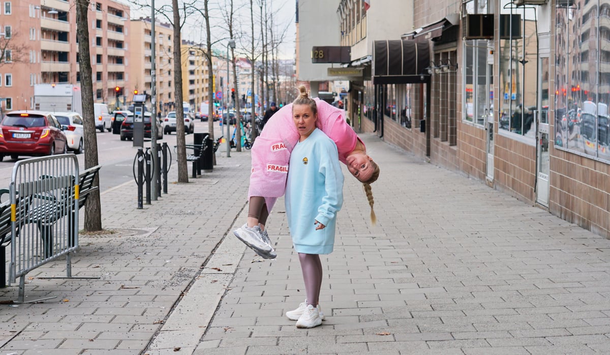 En person bär en annan person över axeln på en trottoar. Båda tittar in i kameran.