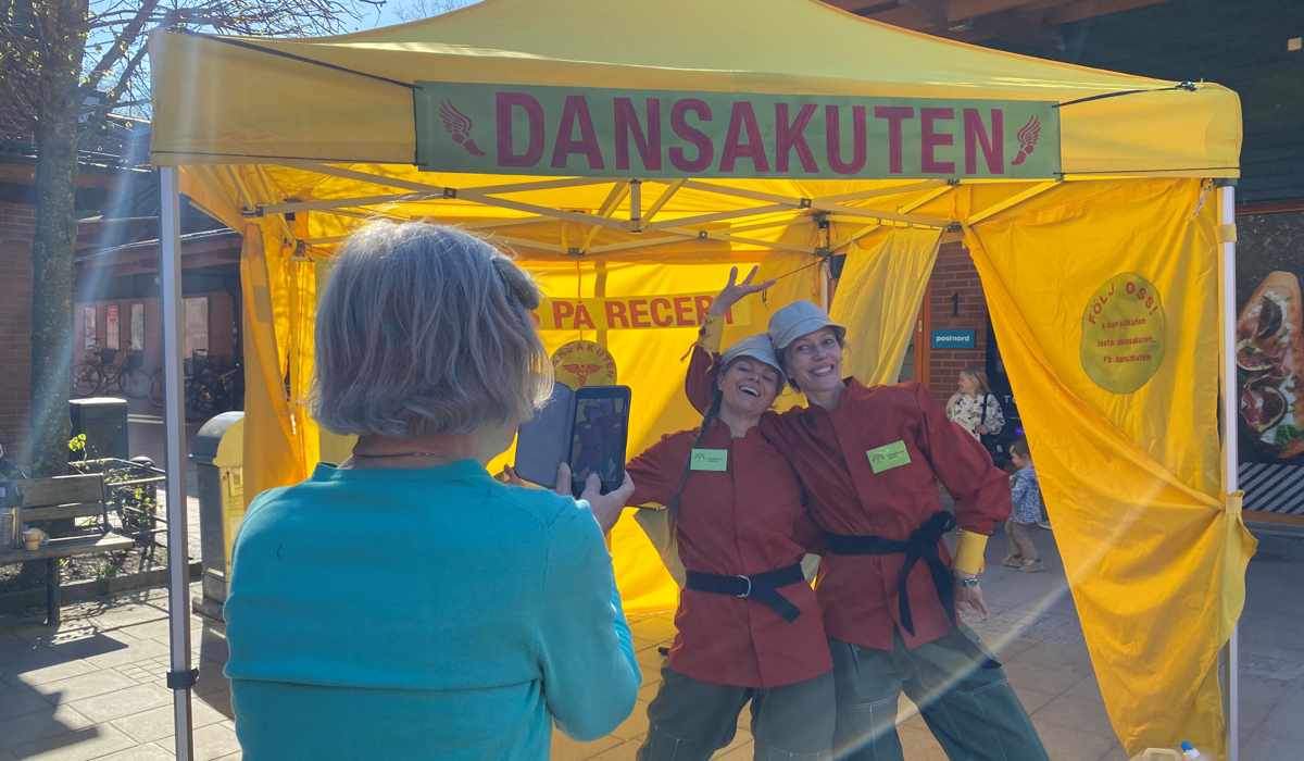 Ett gult partytält på ett torg med texten Dansakuten, två personer i röda jackor och gröna byxor står i tältet och blir fotograferade av en äldre person med mobiltelefon.