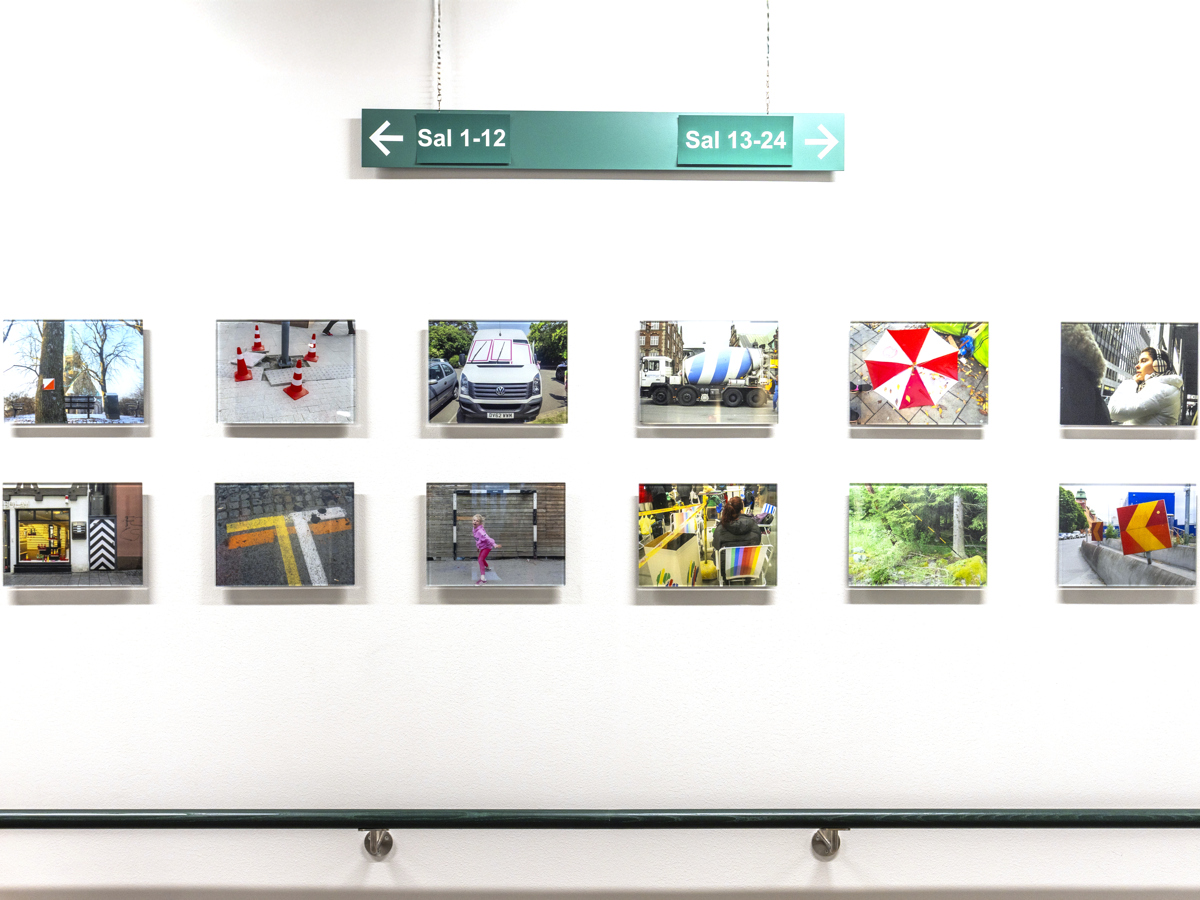 12 små fotografier hänger på en vägg under en sjukhusskylt med text Sal 1-2, Sal 13-24.