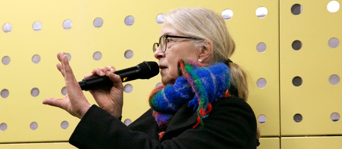 Kvinna med mikrofon framf&ouml;r gul v&auml;gg med h&aring;lm&ouml;nster.