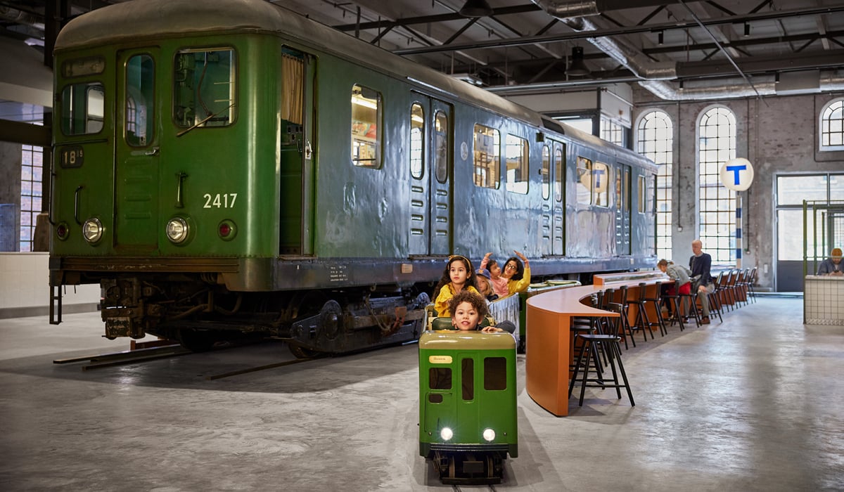 En grupp barn sitter i ett liten tunnelbanevagn, med en stort utst&auml;llningst&aring;g i bakgrunden. 