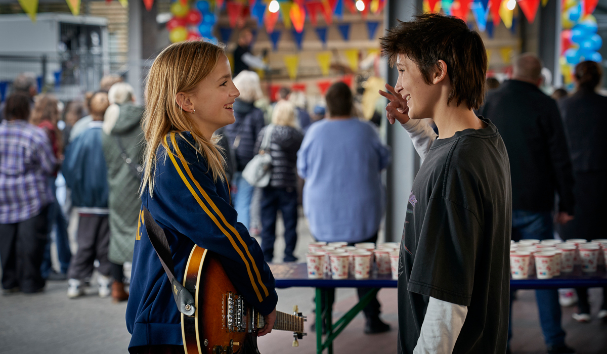En tjej med elgitarr och en kille st&aring;r och pratar p&aring; en plats med partyflaggor och mycket folk