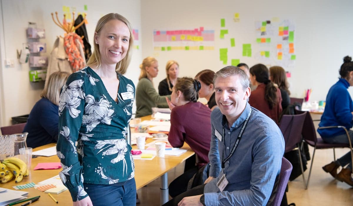 Foto på två personer som ler in i kameran. Den ena står upp och den andra sitter på en stol. I bakgrunden syns andra personer. 