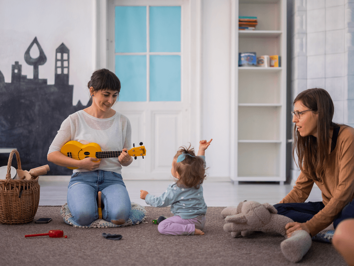 Litet barn på golvet pekar mot himlen. Tittar på kvinna med gitarr. Bakom sitter kvinna och håller nalle.