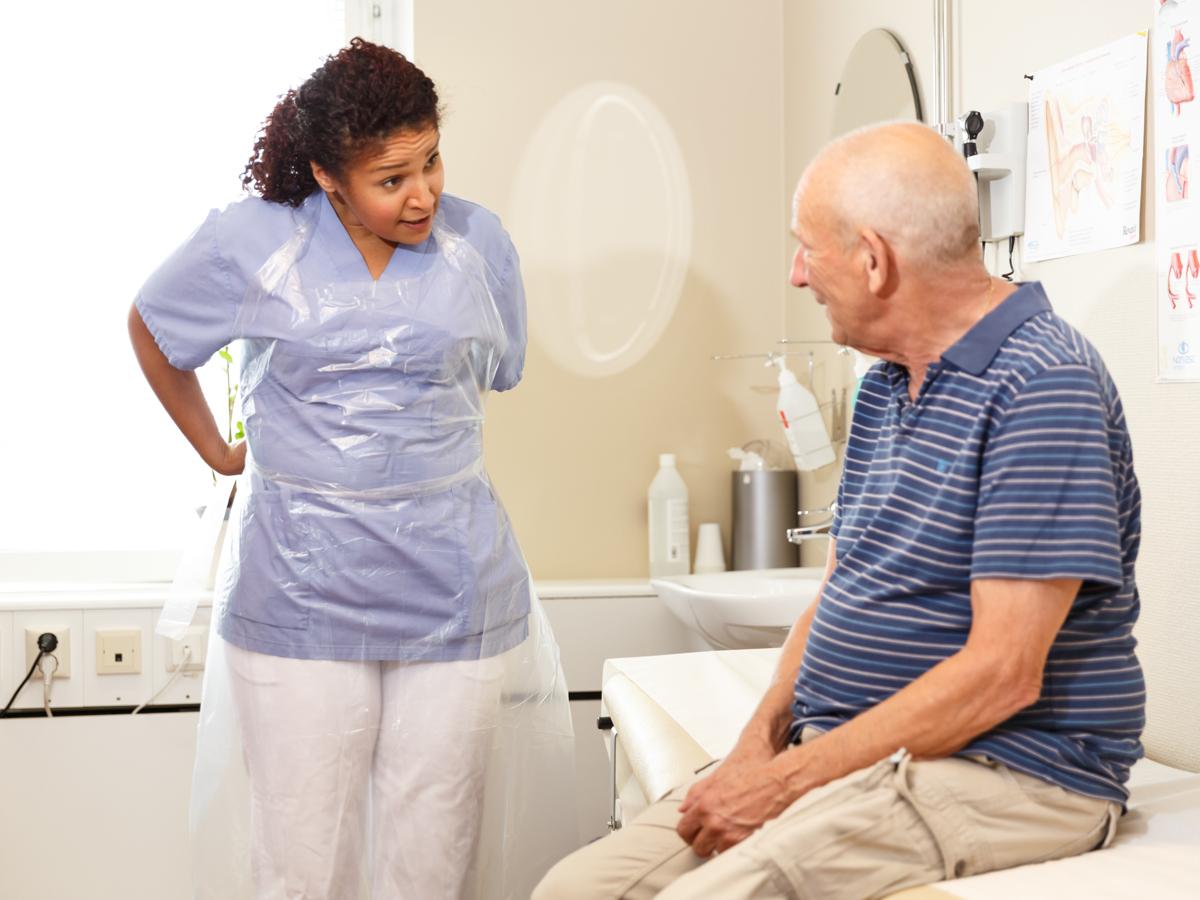 Foto på sjukvårdspersonal som tar på sig ett förkläde samtidigt som hon pratar med en äldre manlig patient som sitter på en brits