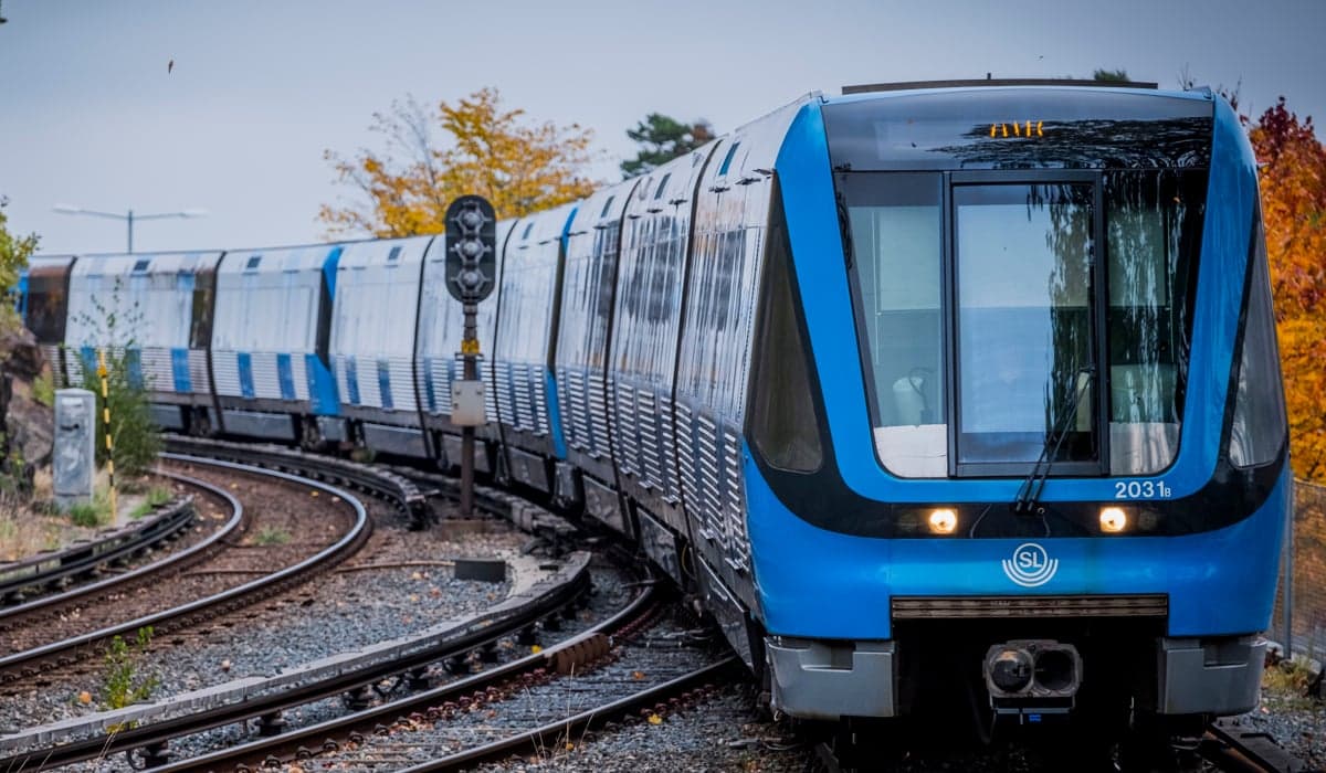 Ett blått tunnelbanetåg rullar in mot station. Runt den så syns träd vars blad är röda och gula, det är tydlig höst. 