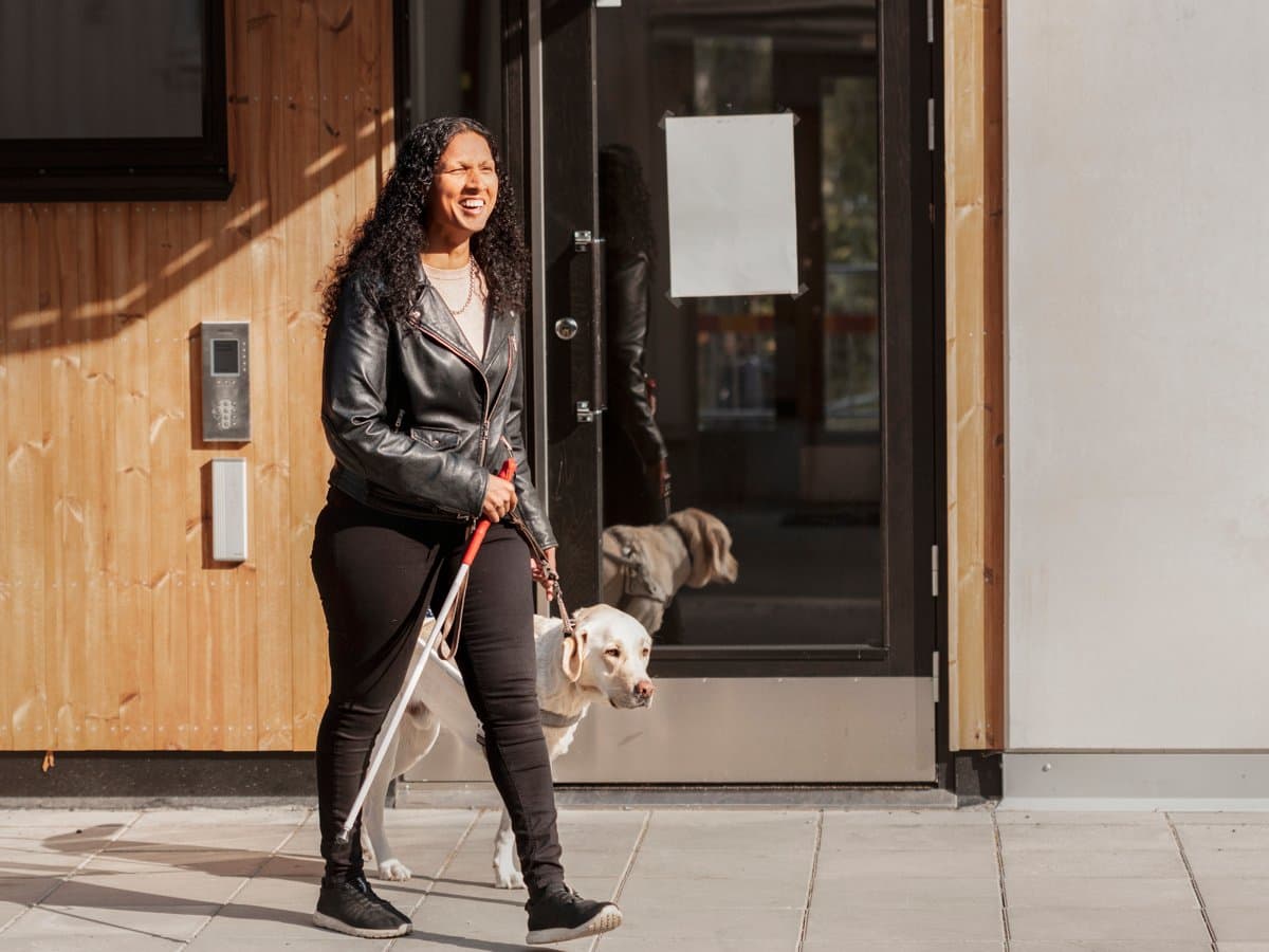 En synskadad kvinna på promenad. I sin ena hand håller hon sin ledarhund, i den andra handen håller kvinnan en blindkäpp