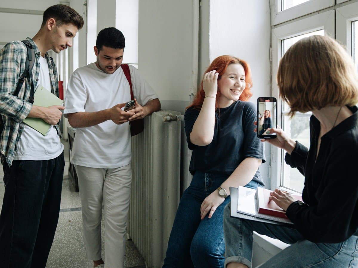 Fyra ungdomar umgås i en korridor. Två pojkar står och tittar på en mobilskärm. Två flickor sitter på en fönsterkarm. Den ena fotar den andra med mobilen. 
