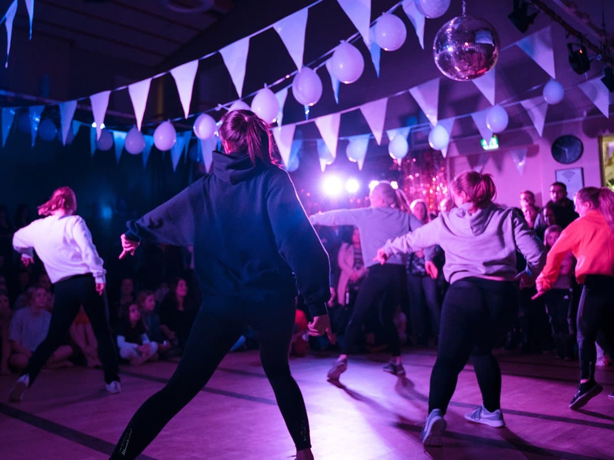 Fem dansare dansar en synkroniserad koreografi på ett dansgolv i en mörk lokal med flaggor, ballonger och festlig belysning i taket.