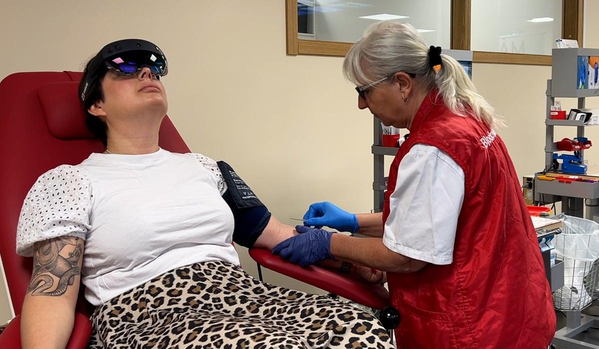 Kvinna  som ger blod ligger på en brits med ett headset som gör att hon samtidigt kan uppleva en animerad värld. En sjuksköterska sätter en nål i kvinnans armveck.
