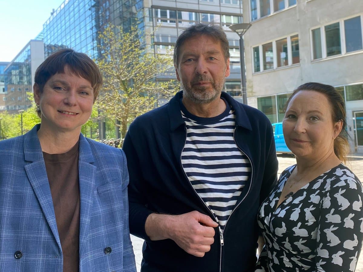 Tre personer står utomhus med hus i bakgrunden