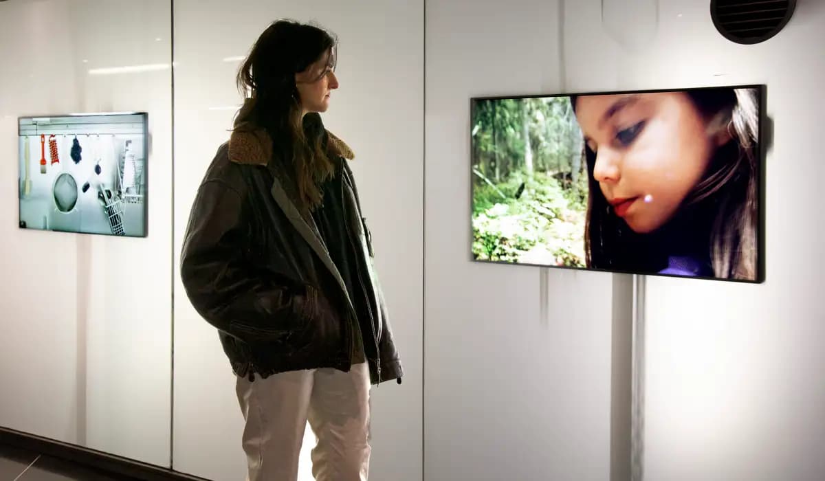 Sophie mellan två av skärmarna på tunnelbanestationen. Hon tittar ned på en av skärmarna där man ser ett närbild av en flickas ansikte som också tittar ned.