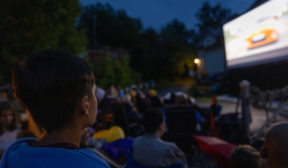 Publik som tittar på bio utomhus. De sitter framför en filmduk, med träd och mörkblå himmel bakom.