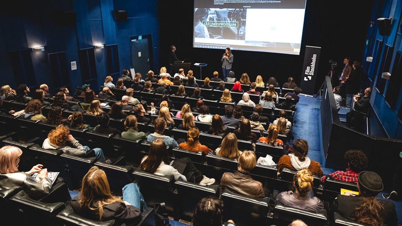 En mörk men fullsatt samlingssal där deltagarna tar del av en presentation och föreläsning