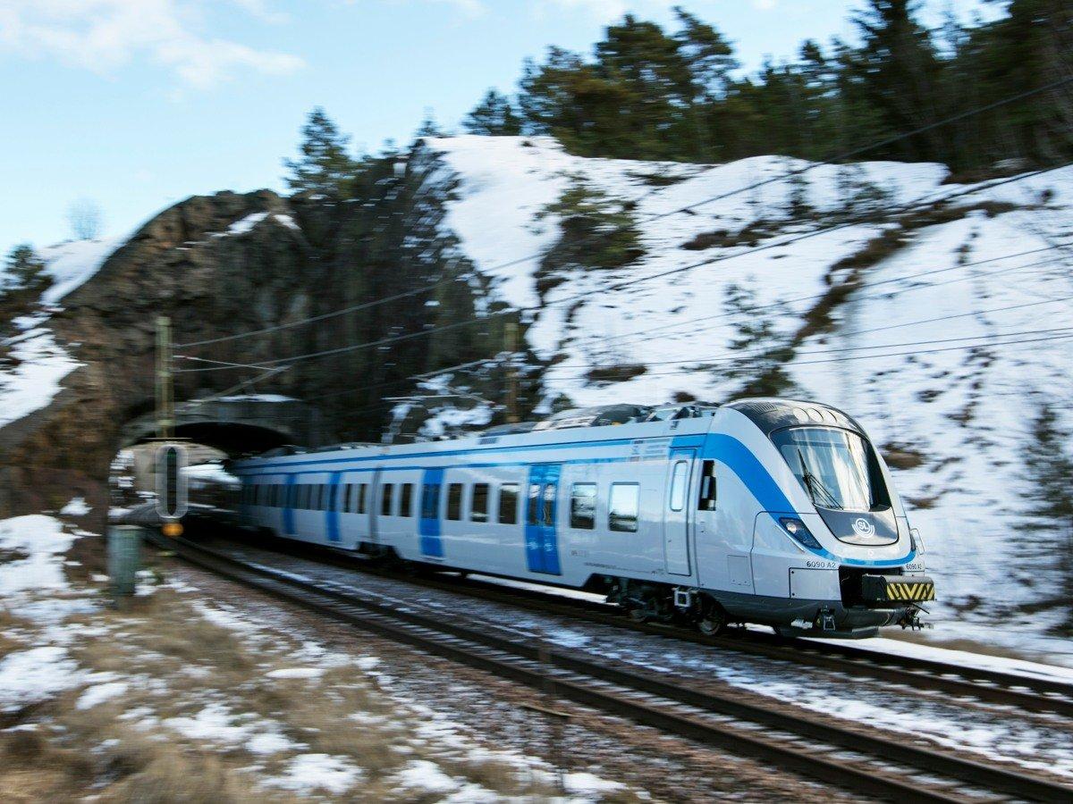 Ett blåvitt pendeltåg åker ut ur en tunnel i hög hastighet. Naturen runtomkring är suddig och bergen är täckta i snö.