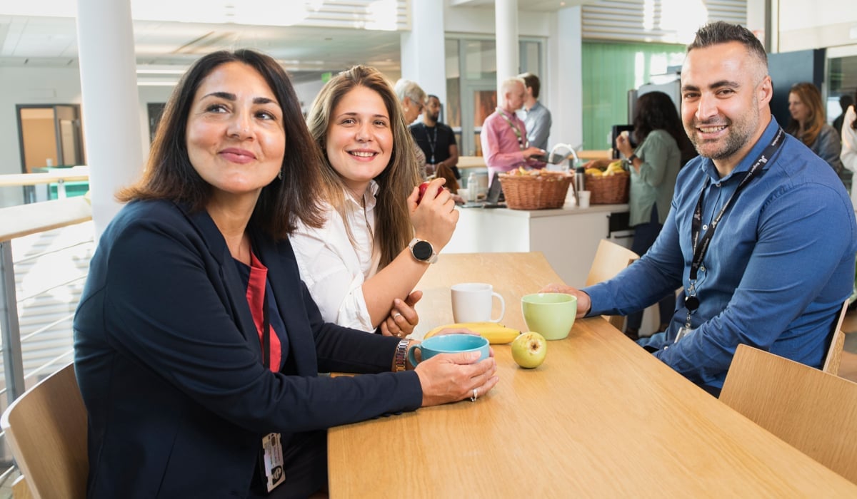 Två kvinnor som sitter bredvid varandra vid ett fikabord amt en man som sitter mittemot. På bordet står det kaffekoppar och ett äpple.