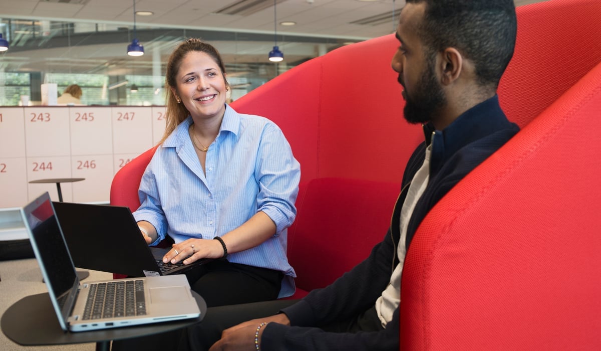 Två medarbetare sitter i en röd soffa och pratar. De har varsin dator.