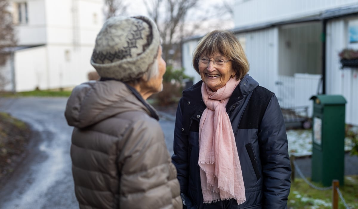 Två äldre kvinnor står och pratar med varandra utomhus. 