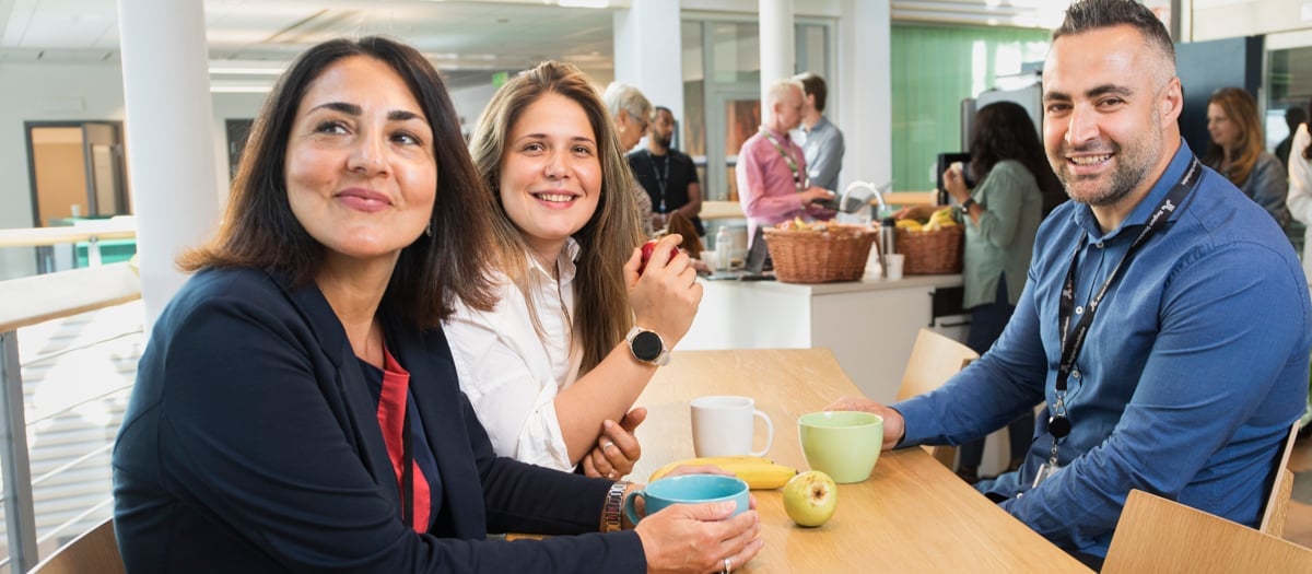 Två kvinnor som sitter bredvid varandra vid ett fikabord amt en man som sitter mittemot. På bordet står det kaffekoppar och ett äpple.