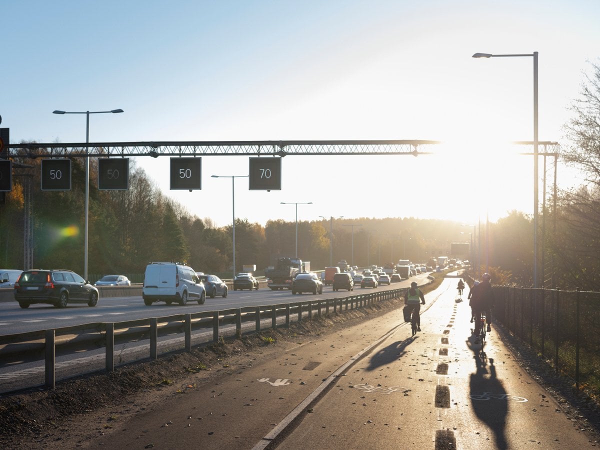 Foto på en bilväg och cykelväg bredvid. På bilden syns bilar och cyklister. 