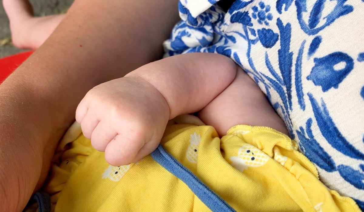 En person håller en bebis i famnen. Bilden fokuserar på bebisens arm och hand.