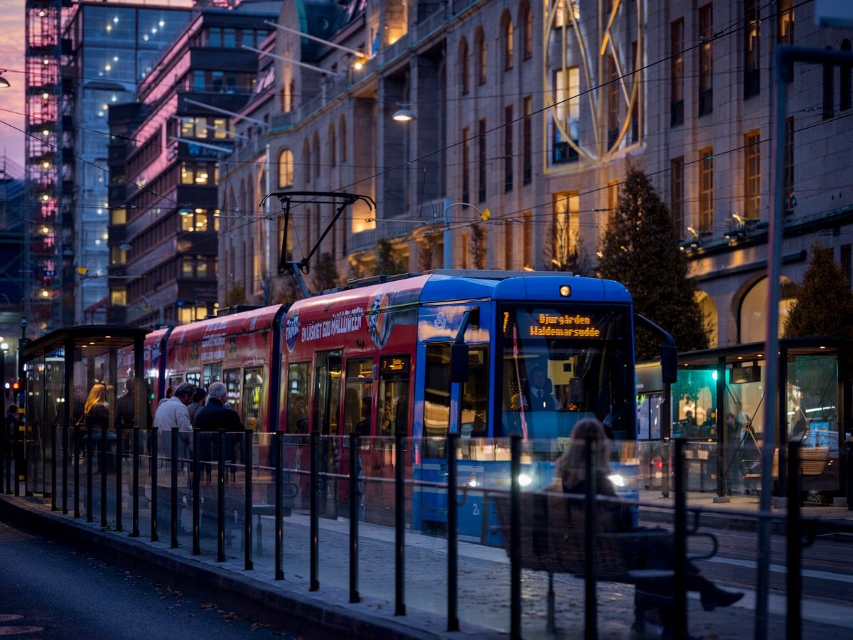 Reklamfoilerad spårvagn stannar vid hållplats med NK-huset i bakgrunden i skymning.