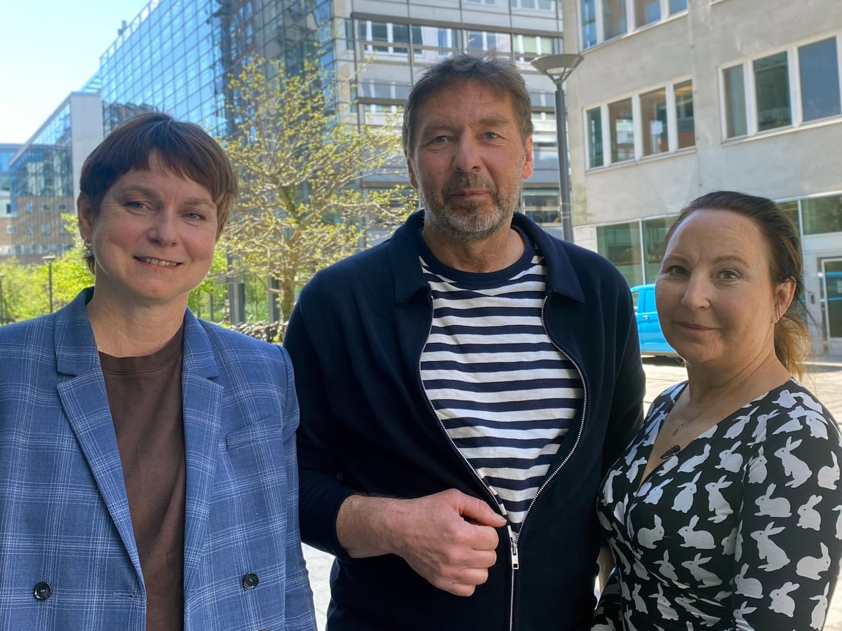 Tre personer står utomhus med hus i bakgrunden