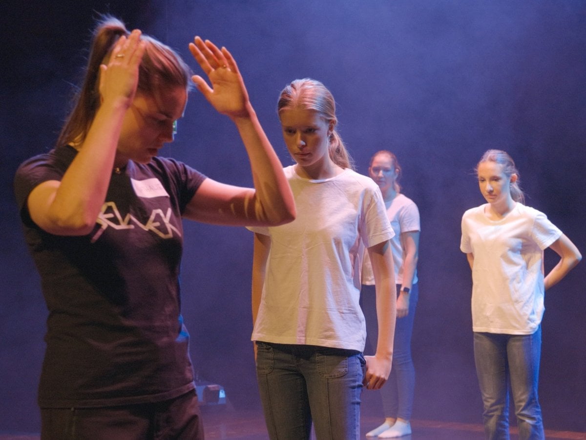 Tre unga kvinnor som är dansare och en kvinna som är koreograf står på ett scengolv och gör rörelser.