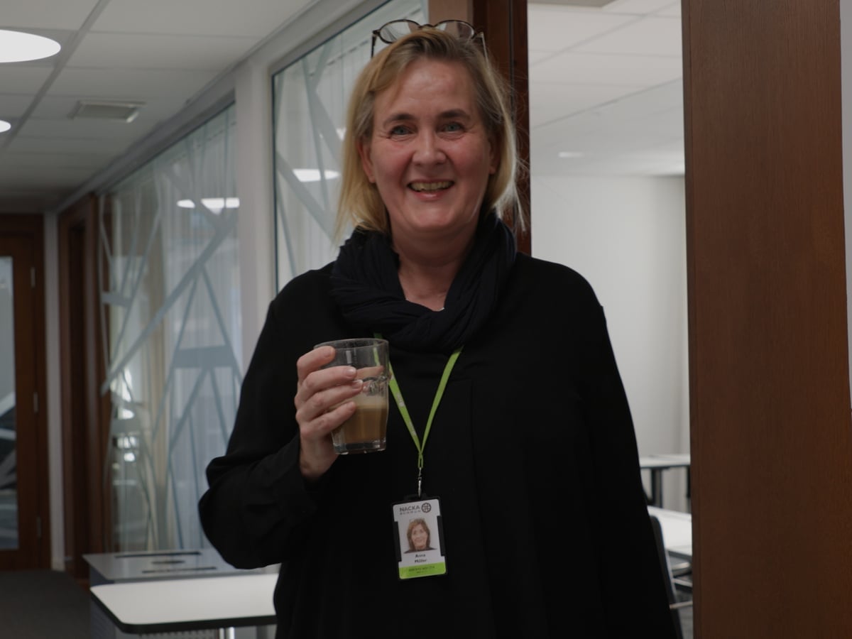 En leende kvinna i en korridor. Hon har ett glas kaffe i handen och ett passerkort i ett band om halsen.