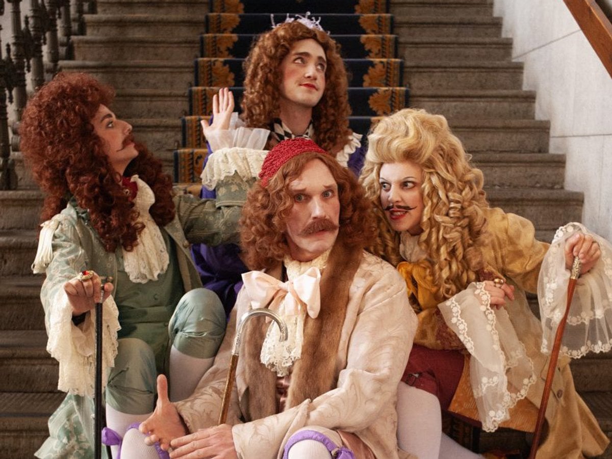 Fyra skådespelare i 1700-talskostymer sitter på en pampig trappa