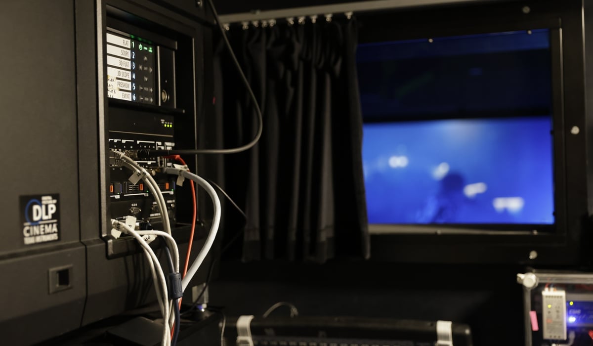 En biografsalong med bioduk sedd från biografens maskinrum. Till vänster i förgrunden syns en digital projektor.