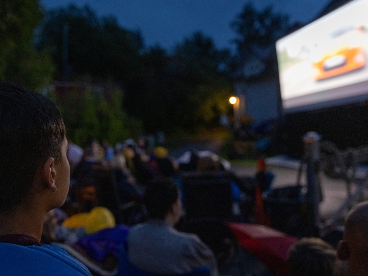 Publik som tittar på bio utomhus. De sitter framför en filmduk, med träd och mörkblå himmel bakom.