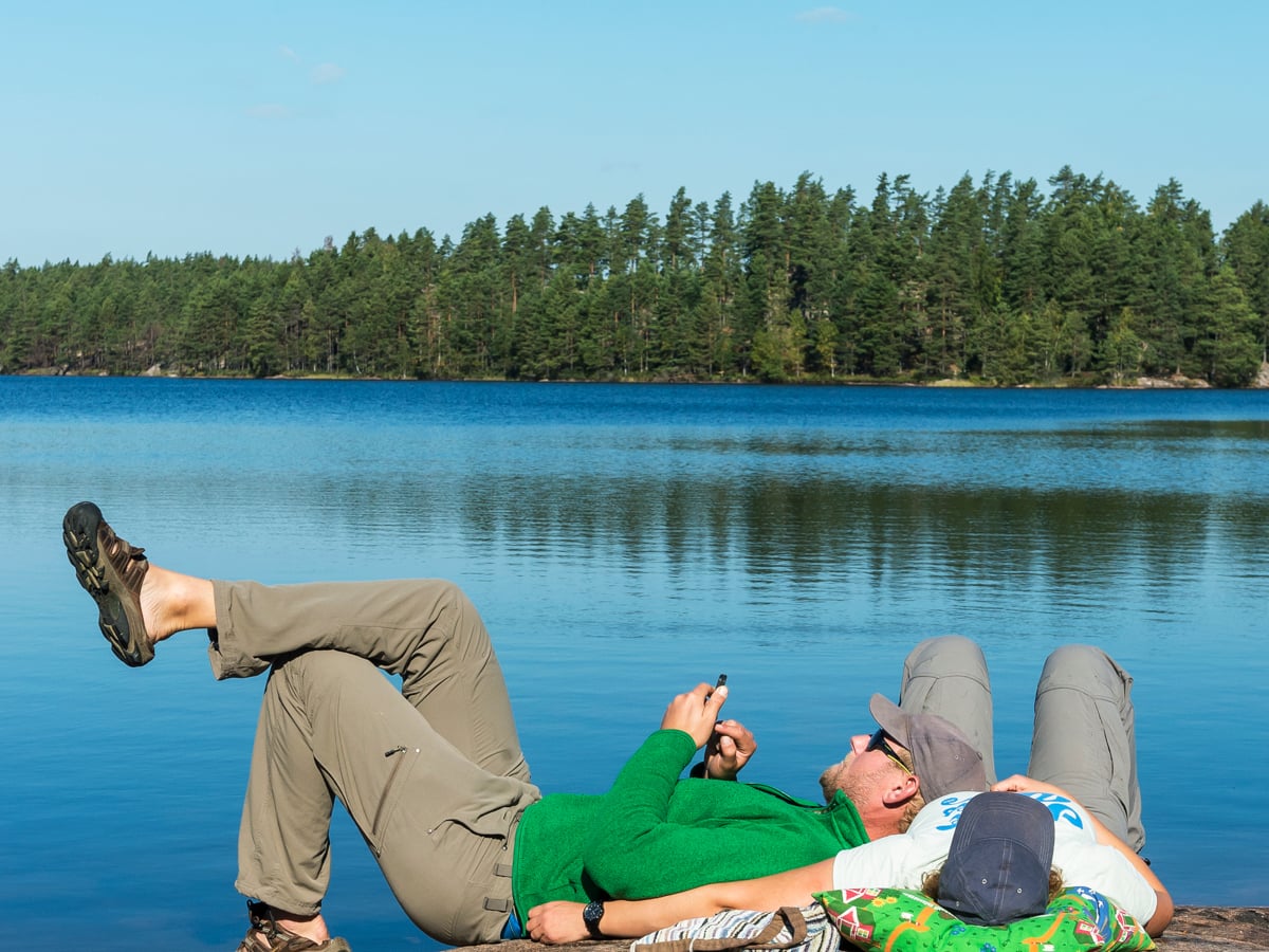 Två personer ligger på en klippa vid vattnet med skog i bakgrunden.