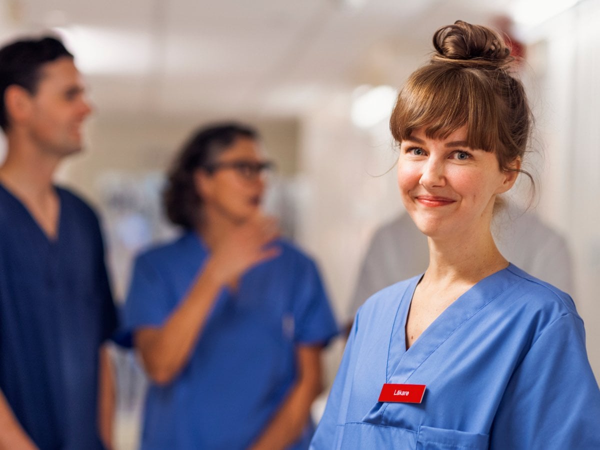 En ung kvinnlig läkare som står i en korridor i sjukhusmiljö. Hon tittar in i kameran och ler. I bakgrunden står tre kollegor till läkaren och pratar med varandra.