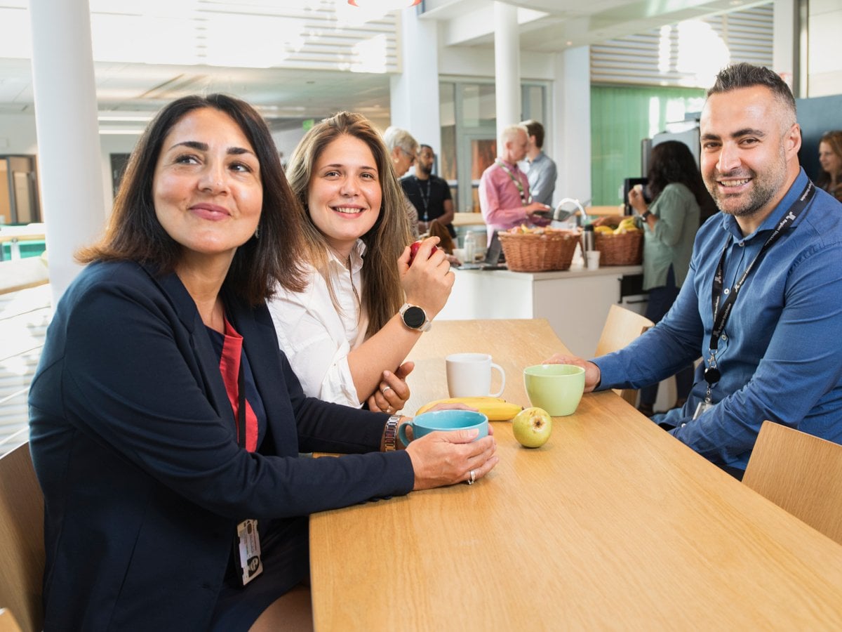 Två kvinnor som sitter bredvid varandra vid ett fikabord amt en man som sitter mittemot. På bordet står det kaffekoppar och ett äpple.