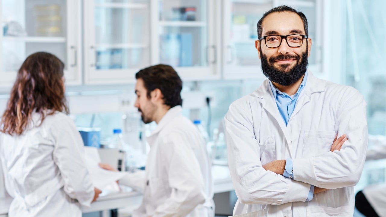 Manlig vårdmedarbetare tittar in i kameran, i bakgrunden ett par kollegor som diskuterar med varandra