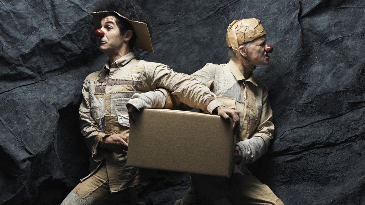 Två personer i beige kläder med röda clownnäsor står med ryggarna mot varandra och håller en brun kartong