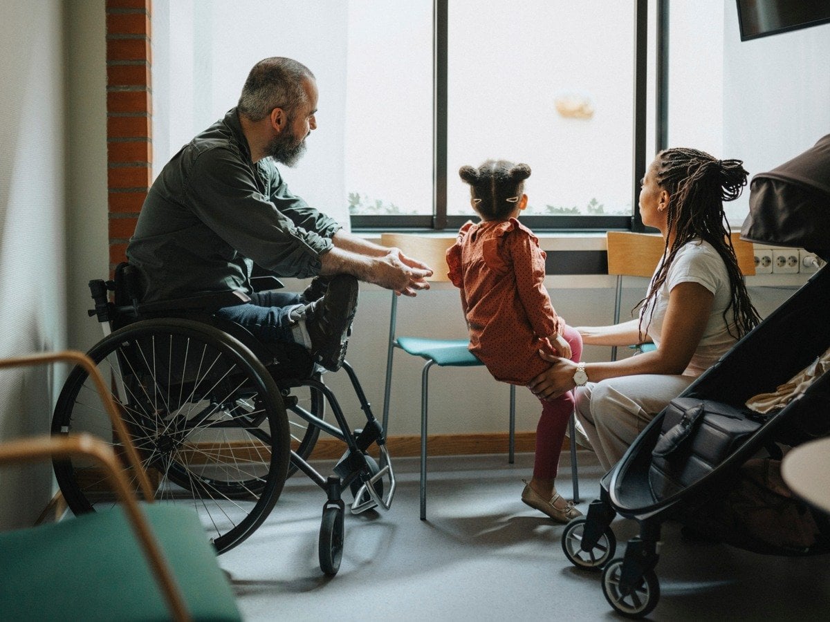 En familj tittar ut genom ett fönster. Pappan sitter i rullstol.