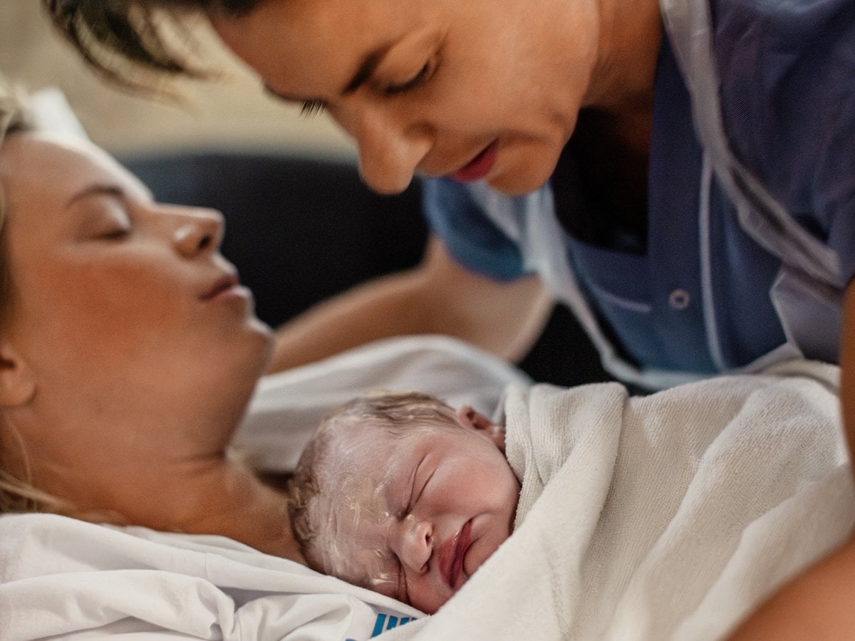Nyfödd bebis som ligger på mammans bröst medan en barnmorska står lutad över.