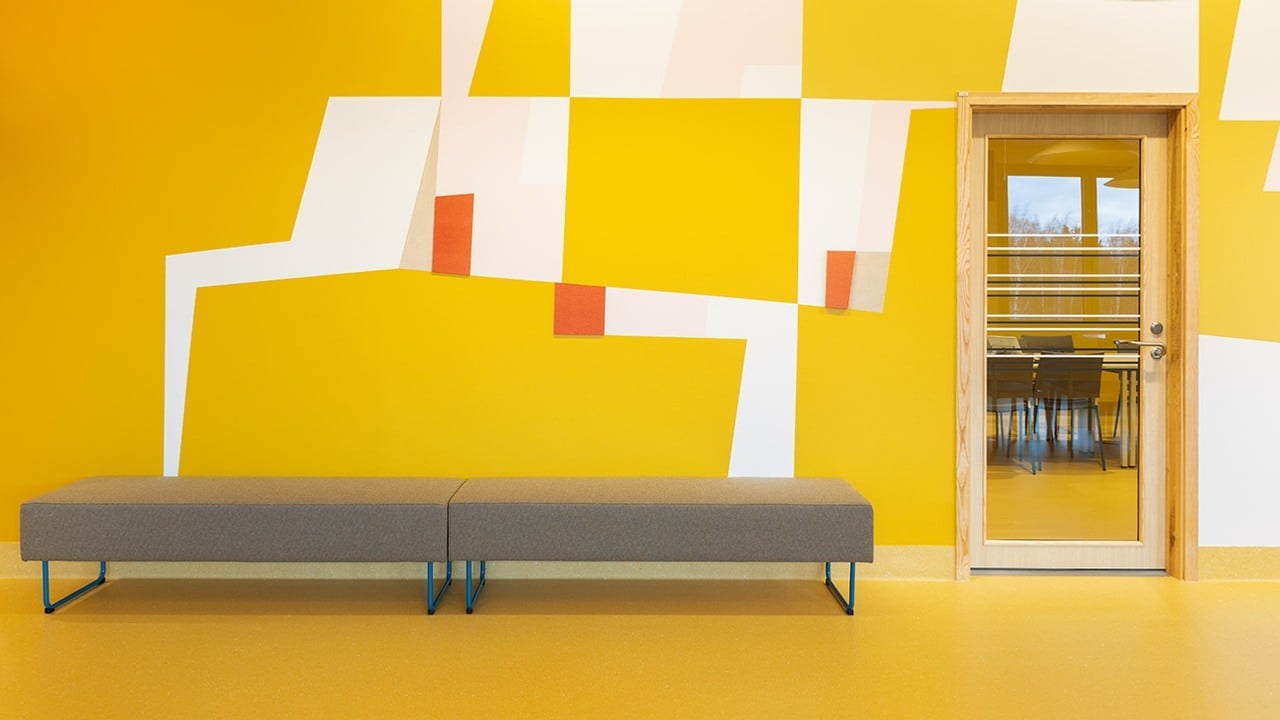 Mönster i gula toner på vägg. Två sittbänkar bredvid glasdörr.