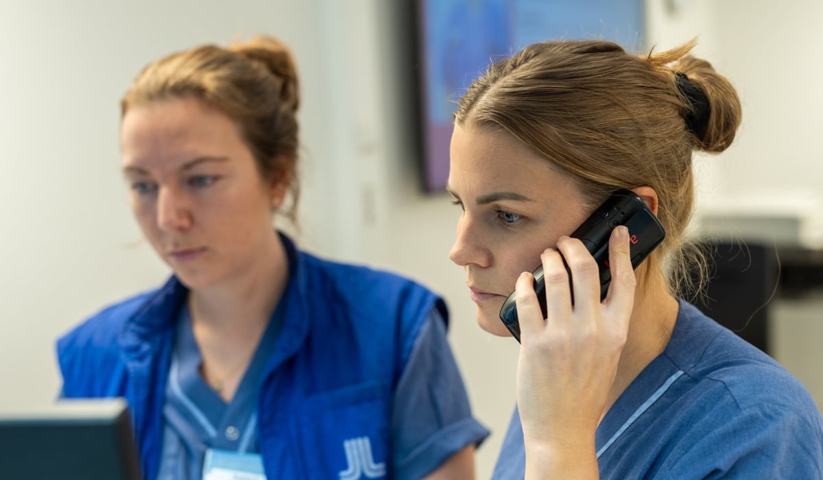 Två sjukvårdspersonal sitter bredvid varandra framför en dator. En pratar i telefon.