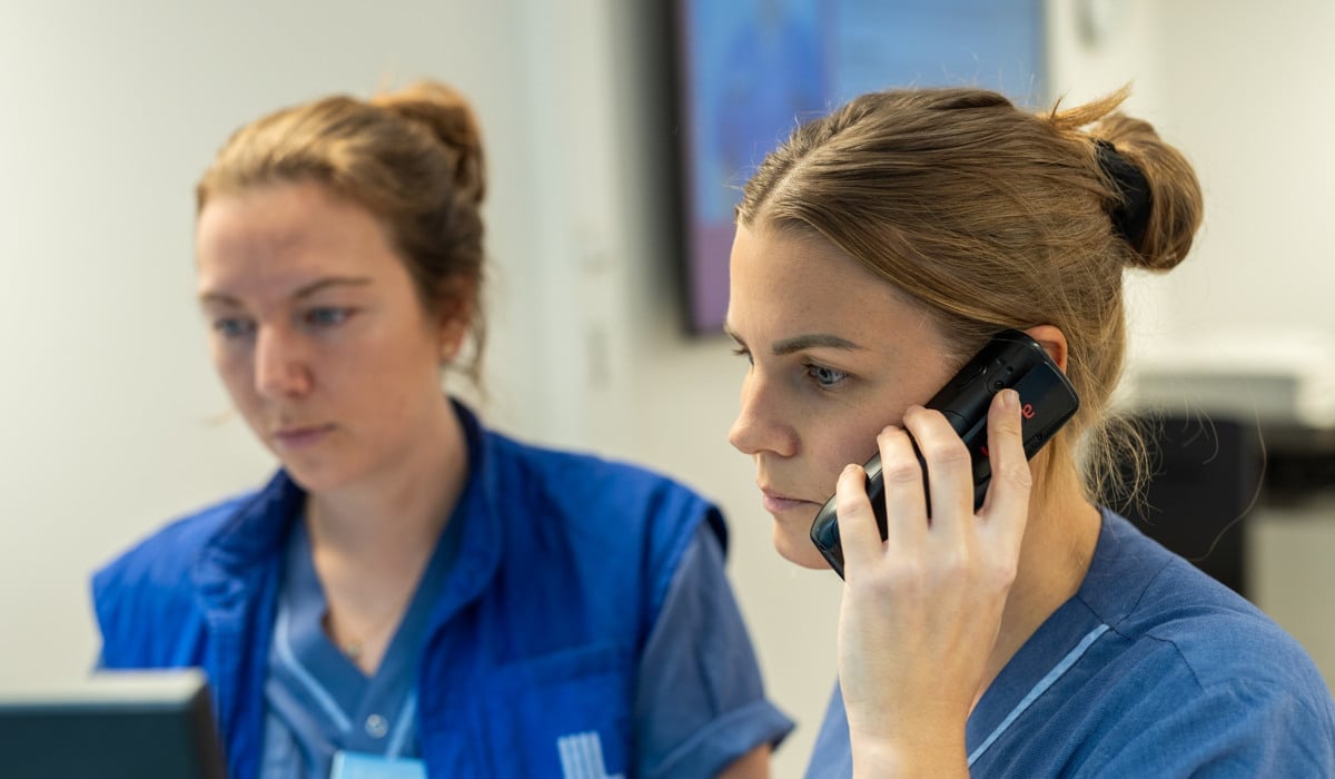 Två vårdpersonal sitter framför en dator. Den ena pratar i telefon.