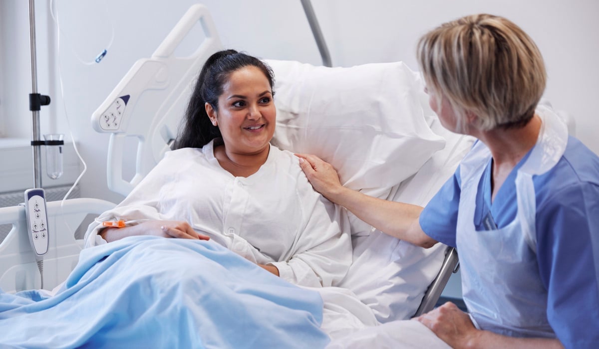 En patient sitter i en sjukhussäng med en vårdpersonal som sitter bredvid och håller handen på hennes axel. 