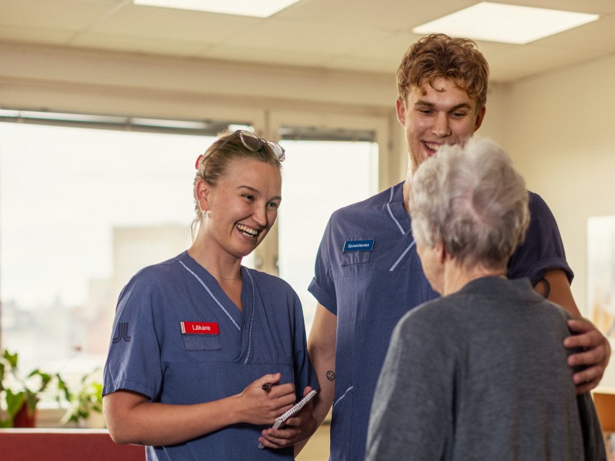 Två personer i sjukvårdskläder står och pratar med en äldre kvinna med rullator.