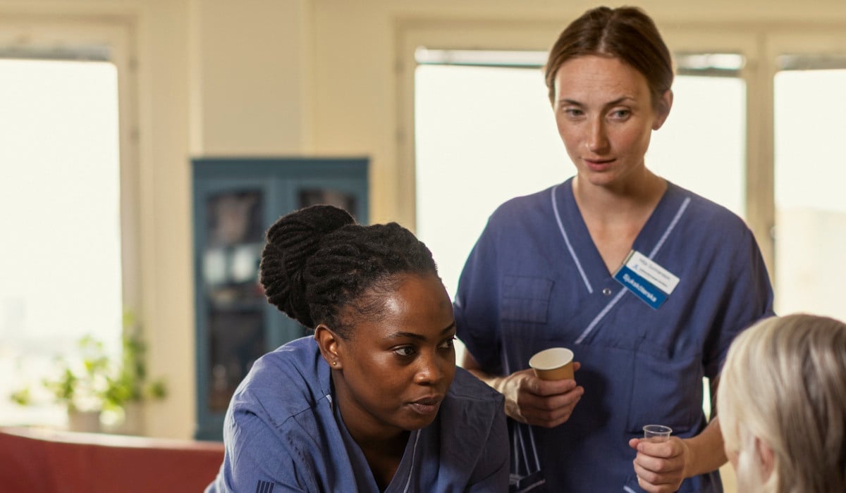 Två personer i sjukhuskläder står och pratar med en person i rullstol.